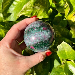 May include: A polished gemstone sphere, primarily green with red and pink inclusions. The sphere is held in a hand with red nail polish, set against a backdrop of green foliage. The gemstone's surface is smooth and reflective.