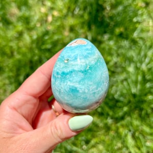 May include: A polished, egg-shaped stone in shades of turquoise and blue, with a small, light brown area at the top. The stone is held against a backdrop of green grass, highlighting its natural colors and textures.