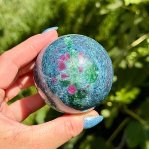 May include: A polished spherical stone featuring a vibrant mix of deep teal and blue base colors, accented with prominent, irregular inclusions of bright magenta-pink and patches of emerald green throughout its smooth, rounded surface.