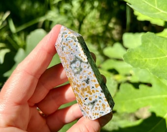 Puede incluir: Una gema rectangular pulida con una parte superior puntiaguda. La piedra presenta un patrón moteado de manchas blancas, verdes y marrón dorado. El cristal se sostiene sobre un fondo de follaje verde.