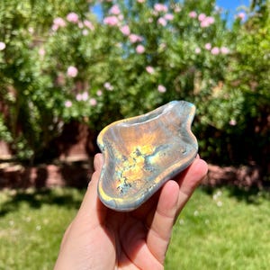 Op de afbeelding: Een handzaam, iriserend labradoriet gua sha-gereedschap. De steen heeft gouden, blauwe en grijze tinten. Het gereedschap wordt in een hand gehouden tegen een achtergrond van groen gras, roze bloemen en een blauwe lucht.