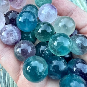 May include: A collection of fluorite spheres in various shades of green, blue, and purple. The spheres are smooth and polished, with a translucent appearance.