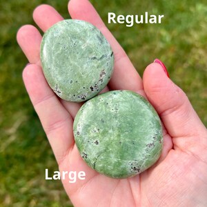 May include: Two green, oval-shaped stones are held in a hand. The top stone is labeled "Regular" and the bottom stone is labeled "Large". The stones have dark spots and a smooth texture. The background is blurred green.