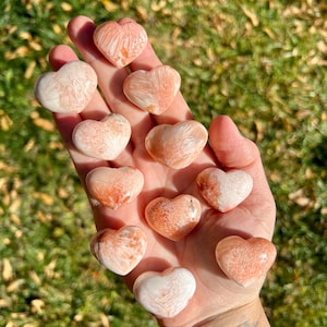 May include: A collection of 12 heart-shaped orange calcite crystals. The crystals are polished and have a smooth, shiny surface. They are arranged in a hand, with the tips of the hearts pointing towards the viewer.