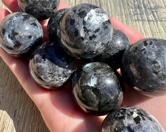 May include: A collection of eight black obsidian spheres, each with a unique pattern of silver flecks. The spheres are smooth and polished, and they are arranged in a hand.