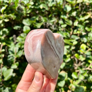 May include: A heart-shaped piece of pink and white banded agate. The agate has a smooth, polished surface and is held in a hand.