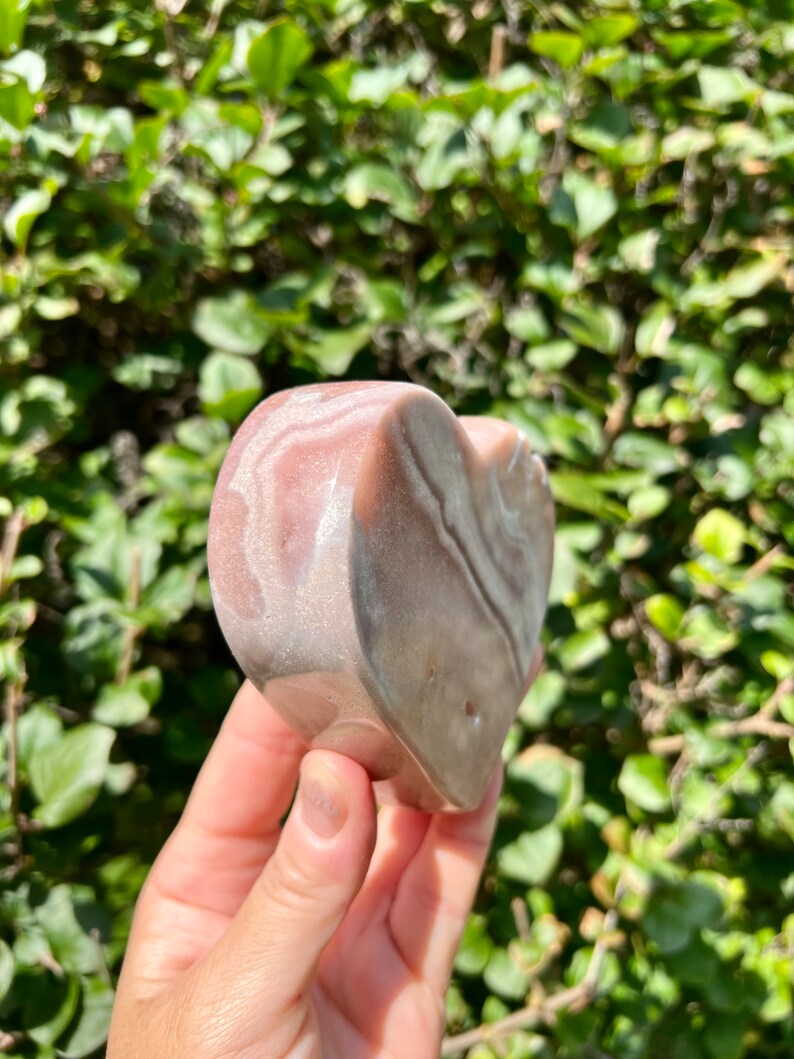 May include: A heart-shaped piece of pink and white banded agate. The agate has a smooth, polished surface and is held in a hand.