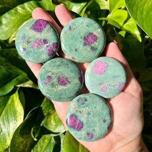 May include: Five round stones with a green base color and vibrant pink and red inclusions are displayed. The stones are smooth and polished, with a natural, earthy appearance. The stones are held in a hand, with green foliage in the background.