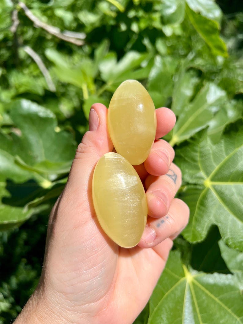 May include: Two yellow oval-shaped polished stones with a smooth, shiny surface. The stones are held in a hand.