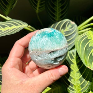 May include: A green and white sphere-shaped crystal with a rough, textured surface. The crystal is being held in a hand.