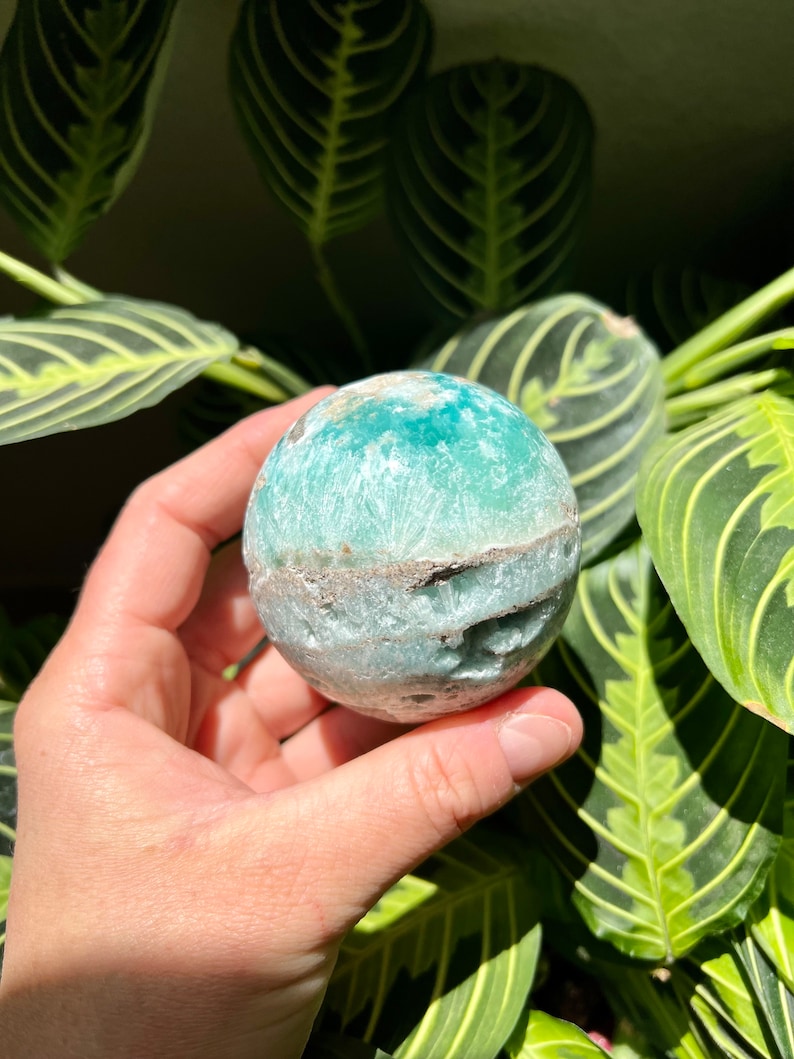 May include: A green and white sphere-shaped crystal with a rough, textured surface. The crystal is being held in a hand.