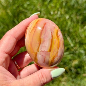 May include: A smooth, oval-shaped stone displaying a mix of pink, yellow, and orange tones. The stone has a polished, glossy finish and a natural, organic look. The colours are arranged in flowing, vertical patterns.