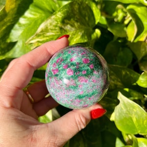 May include: A polished gemstone sphere, predominantly green with vibrant pink and red inclusions. The sphere is held against a backdrop of lush green foliage, creating a natural and appealing contrast. The gemstone's surface reflects light, highlighting its unique patterns.