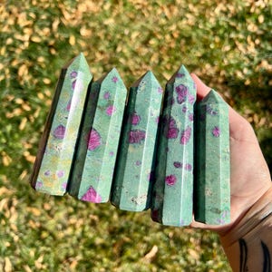 Könnte beinhalten: Fünf grüne Rubin-Zoisit-Kristalltürme mit rosa und roten Einschlüssen. Die Kristalle sind sechseckig und poliert, mit einem natürlichen, erdigen Aussehen. Die Kristalle werden in einer Hand vor einem verschwommenen grün-braunen Hintergrund gehalten.