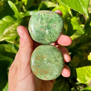 May include: Two round, polished green stones with natural veining and brown inclusions are held in a hand. The stones have a smooth, glossy finish and vary slightly in color, with one appearing lighter than the other. The background is blurred green foliage.