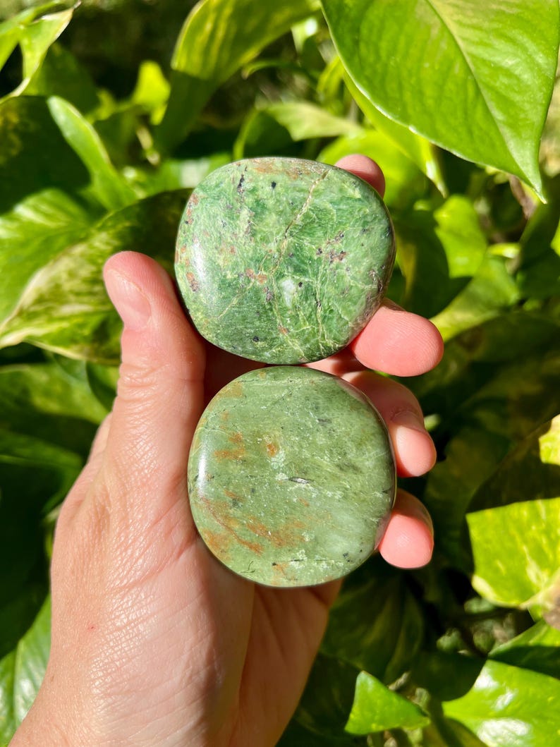 May include: Two round, polished green stones with natural veining and brown inclusions are held in a hand. The stones have a smooth, glossy finish and vary slightly in color, with one appearing lighter than the other. The background is blurred green foliage.