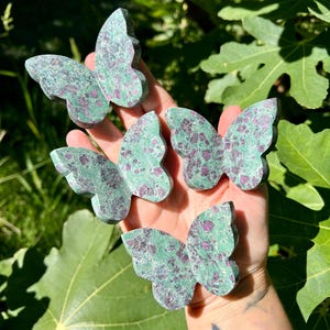 May include: Five butterfly-shaped sculptures. Each butterfly is carved from a stone with a mottled green and red pattern. The butterflies are held in a hand against a backdrop of green leaves and sunlight.