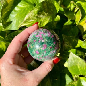 May include: A polished gemstone sphere, primarily green with vibrant pink inclusions. The sphere is held in a hand with red nail polish, set against a backdrop of lush green foliage. A decorative item.