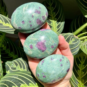 May include: Three smooth, green and purple, polished stones with a speckled pattern. The stones are in the palm of a hand.