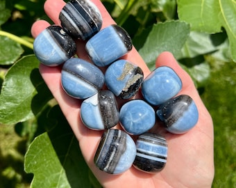 May include: A collection of tumbled stones in shades of blue, black, and white. The stones are smooth and rounded, with varying patterns of stripes and swirls. The stones are held in a hand, with green leaves in the background.