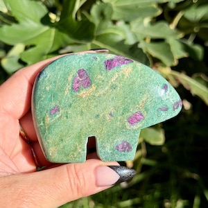 May include: A polished stone carving in the shape of a bear, featuring a vibrant green base color with embedded deep magenta and purple mineral inclusions throughout the surface, showcasing a smooth texture and natural stone patterns.