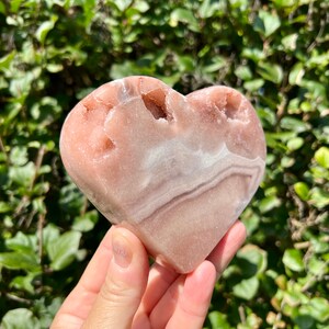 May include: A heart-shaped pink and white crystal with a natural, rough texture. The crystal has a smooth, polished surface and is a natural, organic shape.
