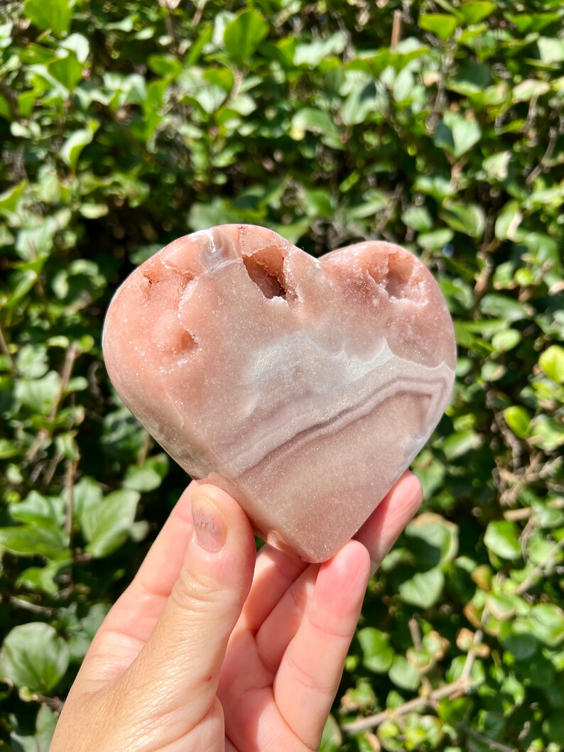 May include: A heart-shaped pink and white crystal with a natural, rough texture. The crystal has a smooth, polished surface and is a natural, organic shape.