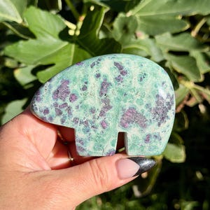 May include: A polished stone carving in the shape of a bear, featuring a mottled light green base color with embedded dark magenta and deep purple mineral inclusions throughout the smooth, rounded surface of the figurine.