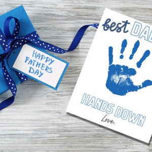May include: A white card with a blue handprint and the text "best DAD ever HANDS DOWN Love."  A blue gift box with a white polka dot ribbon and a tag that says "Happy Fathers Day" is in the background.