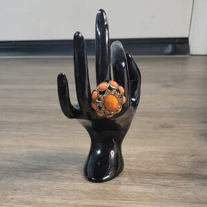 May include: A large, orange statement ring with gold accents and a cluster of smaller orange stones. The ring is displayed on a black ring holder.