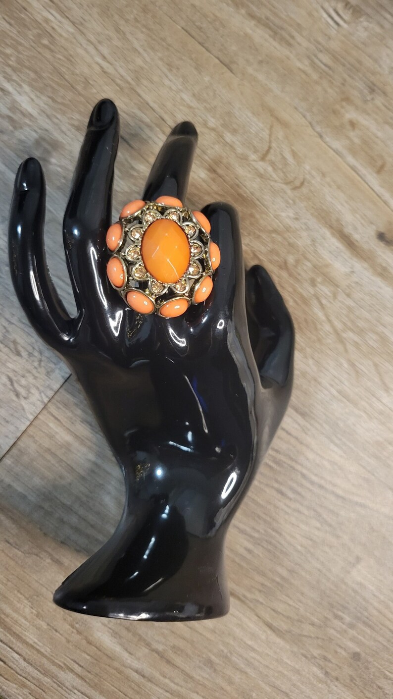 May include: A large, gold-tone ring with an orange oval-shaped stone surrounded by smaller orange stones. The ring is set on a black ring holder.