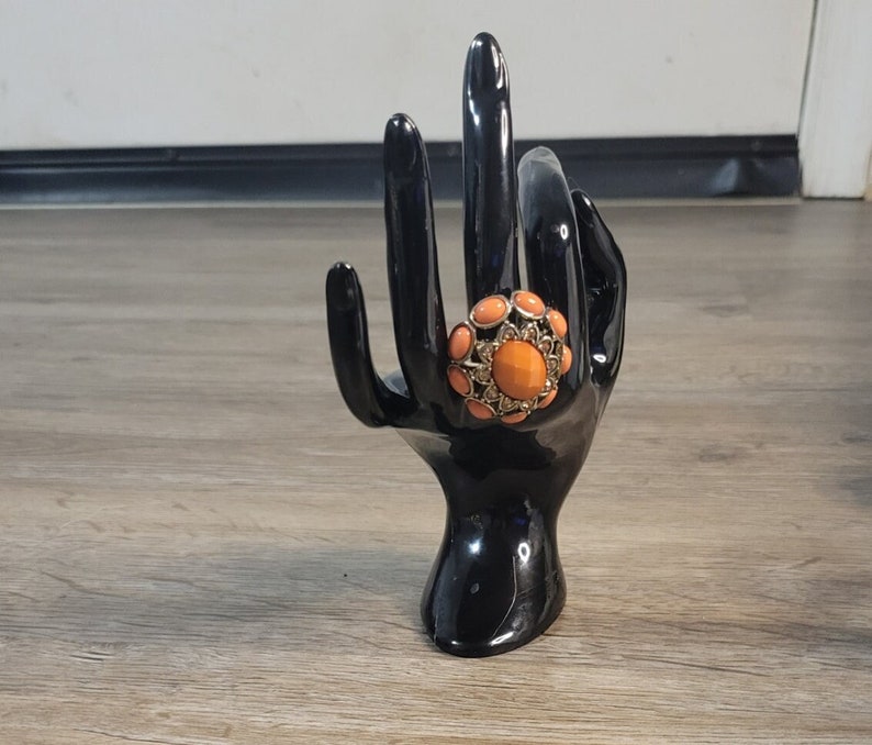 May include: A large, orange statement ring with gold accents and a cluster of smaller orange stones. The ring is displayed on a black ring holder.