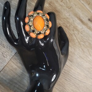 May include: A large, gold-tone ring with a large, oval-shaped, orange stone surrounded by smaller, orange stones. The ring is on a black ring holder.