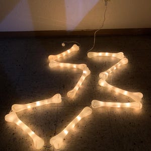 May include: A string of illuminated bone-shaped lights connected by a thin white wire. Each individual bone piece glows with a warm white light, creating a continuous chain of decorative lighting that is plugged into a wall outlet.
