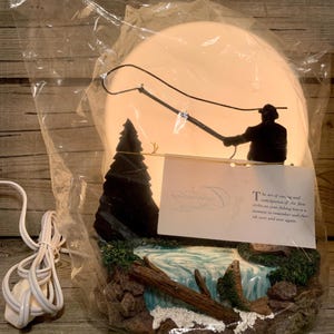 Könnte beinhalten: Dekorative Lampe mit der Silhouette eines Anglers, der seine Angel vor einem leuchtenden Hintergrund auswirft. Der Sockel zeigt eine Wasserfallszene mit einem Fisch, Felsen und einem Baumstamm. Die Lampe ist in Kunststoff verpackt.