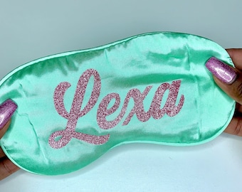 Puede incluir: Una máscara para dormir de satén verde menta con el nombre "Lexa" escrito en purpurina rosa.