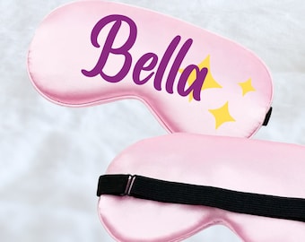 Puede incluir: Máscara de dormir de satén rosa con una correa negra ajustable. La máscara tiene el nombre "Bella" impreso en morado con estrellas amarillas.