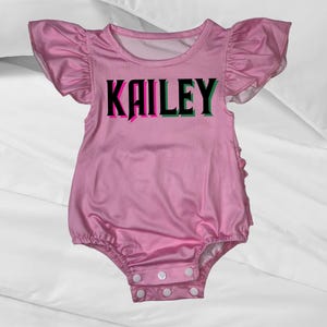 May include: Pink baby onesie with ruffled sleeves and snaps at the bottom. The onesie has the name "KAILEY" printed on the front in black letters with pink and green outlines.