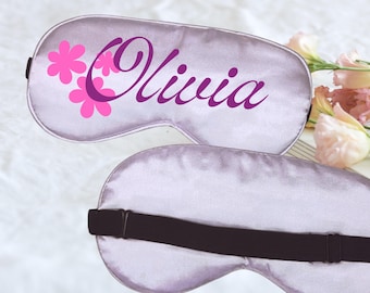 Puede incluir: Dos antifaces de satén lavanda con bandas elásticas negras. Un antifaz presenta el nombre "Olivia" en una fuente cursiva, acompañado de gráficos de flores rosas. El otro antifaz es liso. Los antifaces están diseñados para cubrir los ojos.