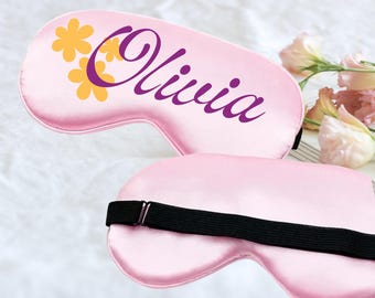 Puede incluir: Dos antifaces de satén rosa. Uno tiene el nombre "Olivia" en escritura morada con detalles florales amarillos y morados. El otro antifaz tiene una banda elástica negra. Los antifaces están sobre una superficie blanca.