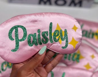 Puede incluir: Máscara para dormir de satén rosa con el nombre "Paisley" en letras de purpurina verde y detalles de estrellas amarillas. Se ven otras máscaras con nombres. Las máscaras son un accesorio para dormir.