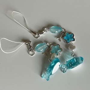 May include: A blue and silver charm bracelet featuring various ocean-themed charms. The bracelet includes translucent blue beads, fish bone charms, star-shaped charms, and two whale charms. The bracelet is attached to a white cord.