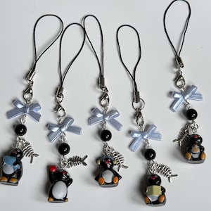 Puede incluir: Colección de cinco colgantes con temática de pingüinos. Cada colgante presenta una pequeña figura de pingüino, una cuenta negra, un lazo azul claro y un dije de espina de pescado plateado. Los colgantes están unidos a cordones negros con cierres plateados.