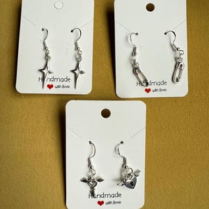Puede incluir: Pendientes plateados con diferentes colgantes: estrellas, clips, una llave alada y un corazón alado. Cada par se muestra en una tarjeta blanca con el texto "Handmade with love" y un corazón rojo.