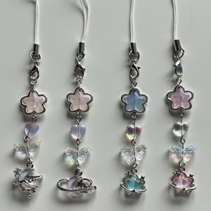Puede incluir: Cuatro colgantes iridiscentes para teléfono con cordones blancos. Cada colgante presenta una cuenta en forma de estrella, cuentas en forma de corazón, dijes de mariposa y un dije con temática celestial. Los dijes son plateados y tienen una estética caprichosa y soñadora.