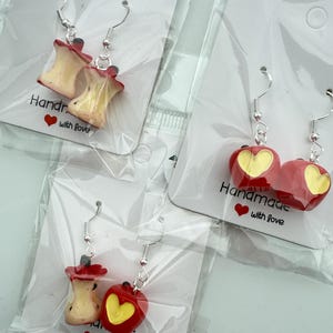 May include: Handmade apple-themed earrings. One pair features red apples with yellow heart cutouts, the other pair shows apple cores. The earrings are on display in clear plastic packaging. The text "Handmade with love" is printed on the packaging.