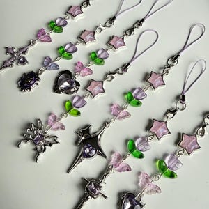 May include: A collection of decorative phone charms with various designs. Charms include crosses, stars, hearts, and floral elements in silver, purple, pink, and green. Each charm has a matching lavender-colored loop for attachment.