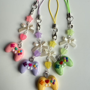 May include: Four colorful game controller charms with pastel pink, purple, yellow, and green controllers. Each charm has a matching colored strap, and is decorated with bows, hearts, and stars. The charms are attached to a silver clasp.