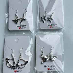 May include: Four pairs of silver-toned earrings, each packaged in a clear plastic bag on a white card. Designs include a heart and key with wings, star shapes, a planet with rings, and a planet with stars. The cards read "Handmade with love."