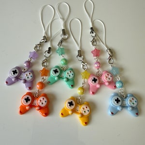 Puede incluir: Una colección de coloridos colgantes de mandos de videojuegos. Cada colgante es de un color diferente, incluyendo morado, naranja, amarillo, verde, rosa y azul. Están adornados con estrellas y cuentas y sujetos a un cordón blanco.